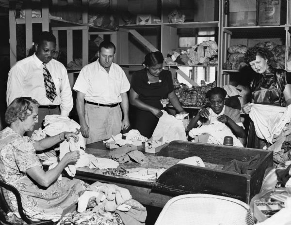 As part of its participation in the local Community Fund, several members of United Packinghouse Workers Local 117 employed by the Kingan Packing Company visited a local Goodwill workshop for handicapped people. The four UPWA workers are (left to right, standing) Ollie Webb, James Biddinger, Josephine Word, and Virginia Burton. Ethel Gray (left) and Edna Crawford are seen at work on a clothing project.