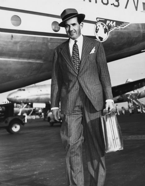 Publicity portrait of CBS news broadcaster Edward R. Murrow. There are airplanes on the tarmac behind him.