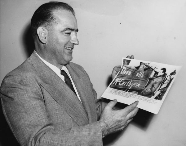 Wisconsin Senator Joseph R. McCarthy laughs in a posed photograph in which he holds a UPI news photograph of a British POW in Korea who denounced his citizenship. He and others are depicted riding in a Korean truck with a banner that says "Down with McCarthyism."