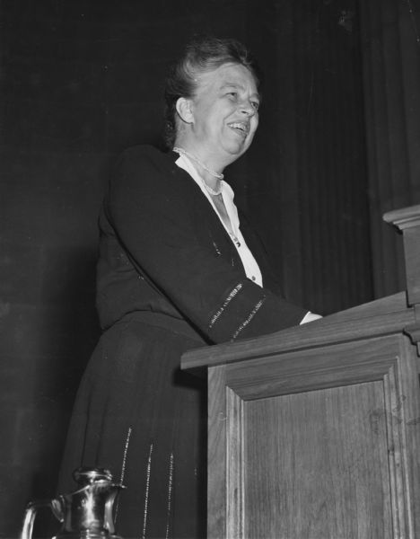 First Lady Eleanor Roosevelt speaking from a podium to an international youth assembly. The original caption indicates that she told the audience that their determination that this should not happen again would go a long way toward preventing another war.