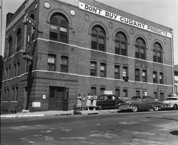 View from across street of various representations of the "Don't Buy Cudahy" boycott campaign outside the Packinghouse Workers headquarters: cartops, women with literature to hand out, and a large banner on the building.