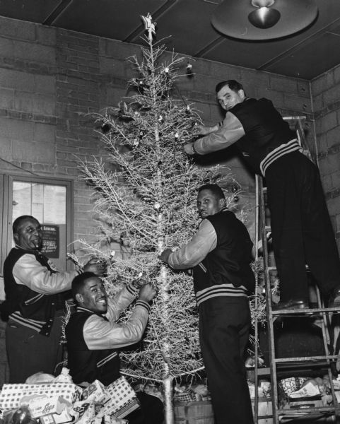 Four members of an unidentified local in United Packinghouse Workers of America District 1 (Burette King, Sidney Coleman, Aljay Williams, and Joseph Kuzma) trim an artificial Christmas tree. Below the tree are baskets of food.