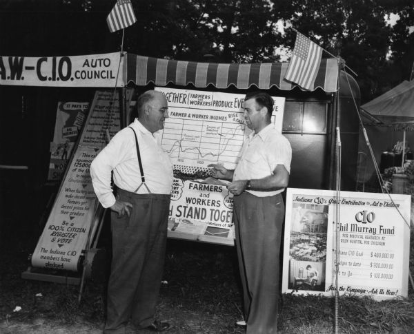 Two men stand in front of a booth sponsored by the Congress of Industrial Organizations at a fair. In addition to displays on farmer-labor solidarity and voter registration, the booth includes a display on funds the union was raising to honor Philip Murray.
