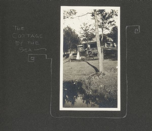View of the Drew family cottage on Lake Flora Dell. A caption of "The Cottage by the Sea" is to the left of the photograph, in addition to a decorative border. A girl is standing in the yard, and a man is on the porch.
