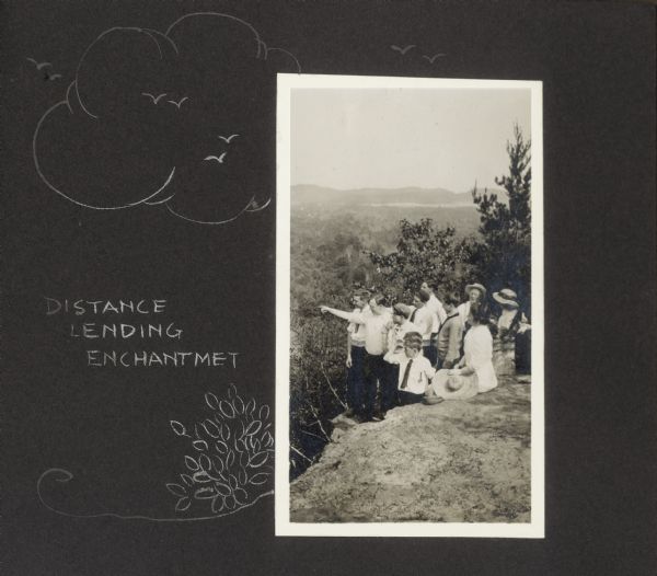 A group of individuals are gathered on a bluff. One man is pointing off to the left, and the rest of the group gazes in that direction. Features a hand-drawn decorative border with a caption that reads, "Distance leading enchantment." The man pointing is identified as Walter Drew, third from left Frank O. King, and seated on a rock wearing a white dress is Delia King. On the far right wearing the banded hat is Doris Drew.