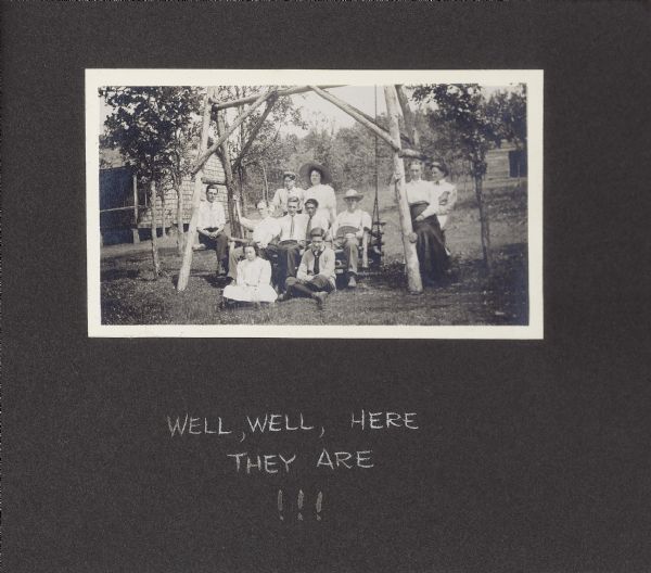 A group of individuals pose for a photograph while seated on and around a log lawn swing in the yard of the Drew cottage. Caption reads, "Well, well, here they are!!!" Individuals are identified as: seated left on swing, Walter Drew; seated right on swing, Frank Drew; standing behind swing, right, Delia Drew; sitting on ground, left, Doris Drew; and far right, leaning right, Lillian Drew.