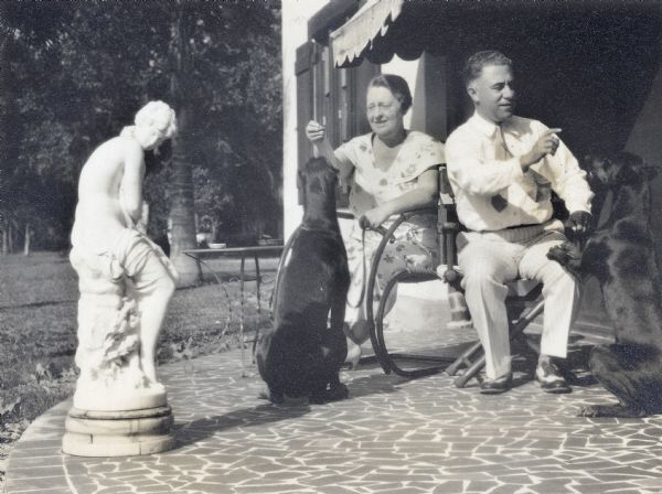 Delia and Frank King on a terrace on the side of a house with their Doberman Pinschers at Folly Farm. A classical sculpture is in the foreground.