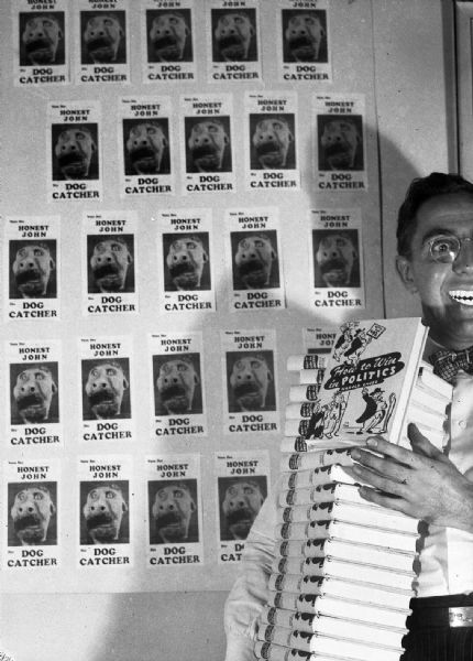 Humorous photograph of Gauer hawking his just published book: "How to Win in Politics." He is standing in front of a wall with multiple copies of a small satirical political flyer for "Honest John / Dog Catcher."