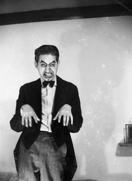 Robert Bloch wearing make-up makes a frightening face while holding his hands out in front of him. His hair is slicked back and he wears a suit jacket with tails and a bow-tie.