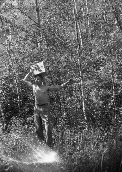 Harold Gauer, smoking a pipe, stands shirtless in a clearing surrounded by trees holding a CARE box on top of his head, labeled: "CARE Send a Care Package to Yugoslavia."