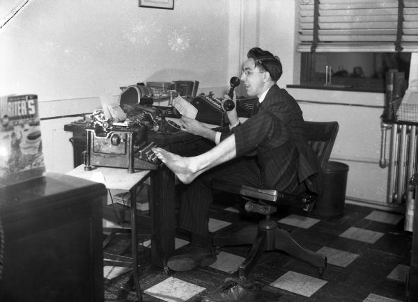 Harold Gauer is sitting at a desk wearing a pinstriped suit. His left foot is bare and "typing" at a typewriter, while he reads a note and talks on the telephone.