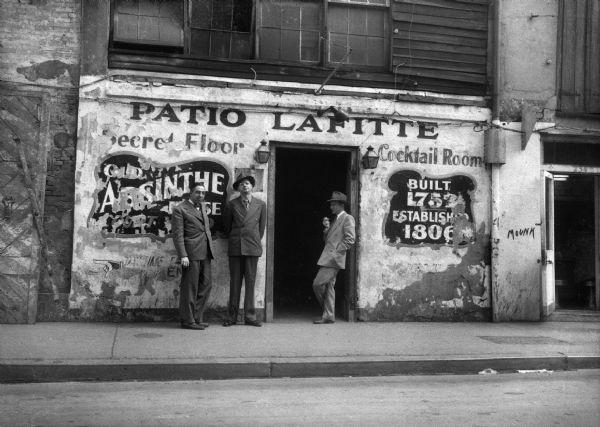 Harold Gauer, Robert Bloch, and an unidentified man holding a cigarette, stand in front of an open doorway in a weathered building. Around the entrance is painted the words: "Patio Lafitte," "Secret Floor," "Cocktail Room," "Old Absinthe Court," and "Built 1752 Established 1806."