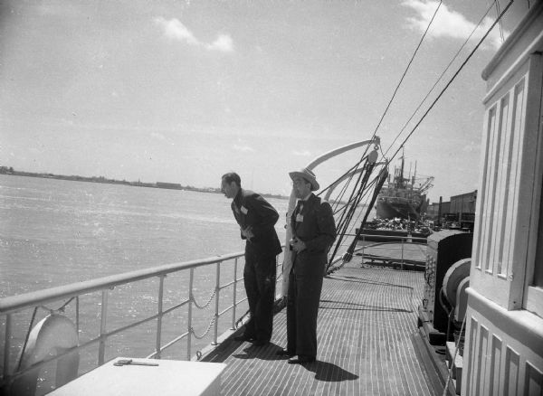 Harold Gauer and Robert Bloch on the deck of a boat near the railing acting as if they were seasick. In the background is another ship tied up at a dock, and in the far distance a shoreline.
