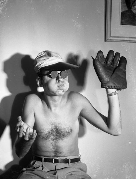 A shirtless Robert Bloch is posed sitting puffing out his cheeks. He is wearing a baseball cap, a baseball mitt, and eyeglasses with covered lenses.