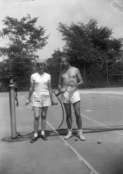 Harold Gauer stands with an unidentified woman in front of a tennis net, both holding tennis racquets.