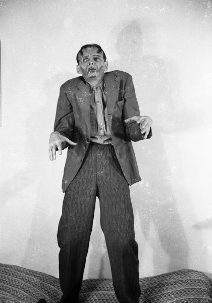 Robert Bloch stands posing on a bed with his hands out in front of him and with monster make-up on his face. He is wearing a jacket, shirt, and long pants.