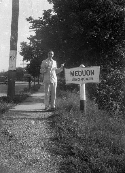 Harold Gauer, wearing a suit and bow tie, and holding an ice cream cone, stands smiling next to a sign on the shoulder of a road that reads "Mequon / Unincorporated."