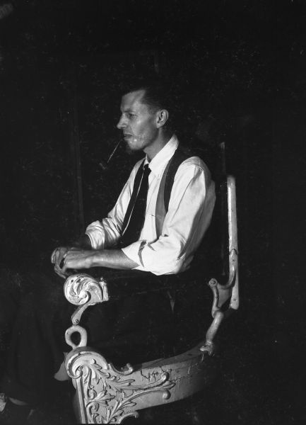 Side view of a man with a goatee smoking a pipe while sitting in a carved chair made of iron and wood.