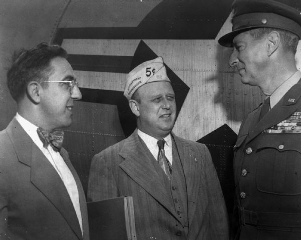 Harold Gauer, on left wearing a bow tie, with two unidentified military officers, one of whom is in full decorated uniform, in front of an airplane.