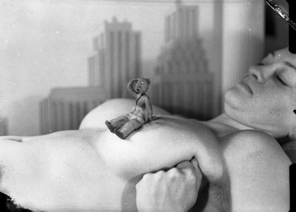 A tiny clay figure sits on the exposed left breast of Alice Bedard, looking into the distance. A poster of skyscrapers is visible in the background.