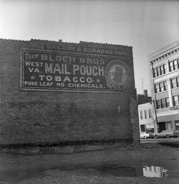 View across empty lot of an advertisement painted on the side of a brick wall, reading: "Chew Bloch Bros' West VA. Mail Pouch Tobacco Pure Leaf No Chemicals."
