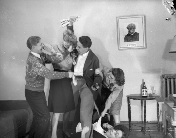Angie Vail and Alice Bedard pose in an attempt to break up a staged fight between Robert Vail and Sprague Vonier, while Harold Gauer lies on the ground underneath the scuffle. There is a bottle of "Beech Rye Whisky" on the table next to the fight, and a framed picture on the wall, presumably made by Gauer, reads "Prexy."
