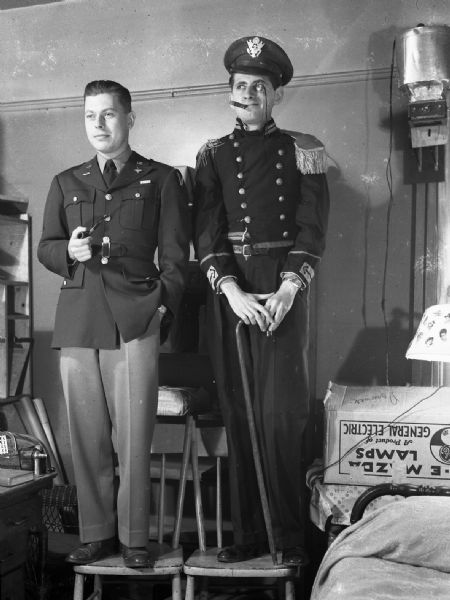 Robert Bloch, with cigar, and Robert Vail, holding a pipe. They are both wearing military costumes, staged standing on chairs next to a bed.