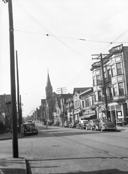 View from sidewalk of cars parked along Brady Street. Storefronts and a church are visible.