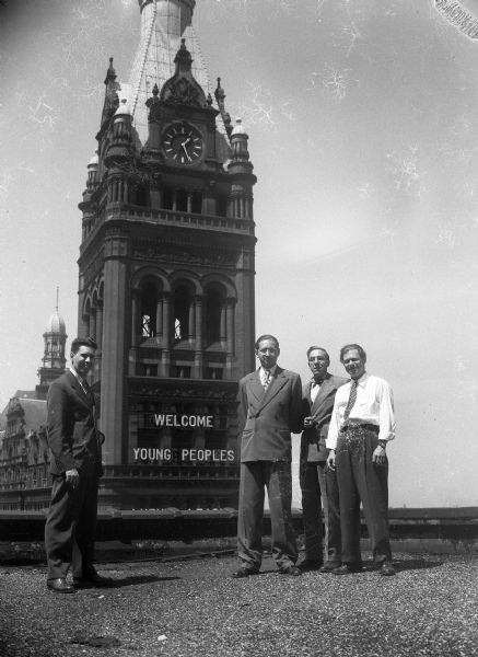 From lef to right, Sprague Vonier, Robert Bloch, Harold Gauer, and Robert Vail stand on the roof of a building. Behind them is a clock tower with the words "Welcome Young Peoples" on it.