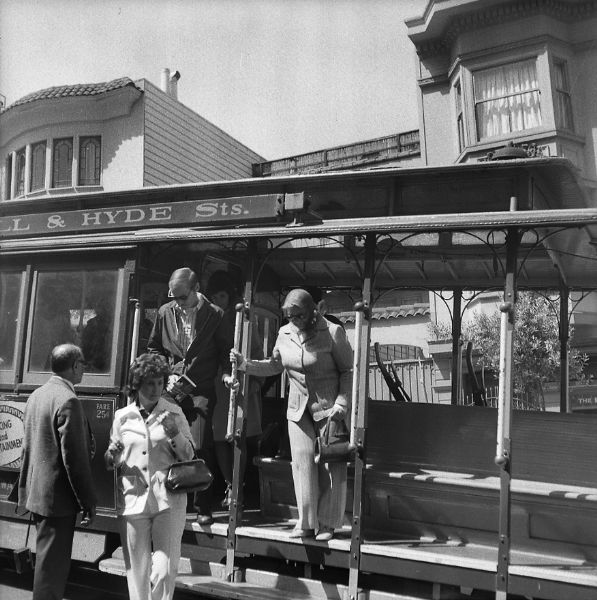 A group of unidentified people getting off of a trolley with the words [obscured]LL & HYDE Sts. on the side of the roof.