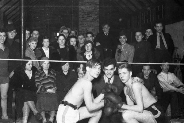 Two young boxers posing kneeling in a ring touching gloves, while the referee crouches between them. An audience of unidentified youths are sitting and standing behind the rope.