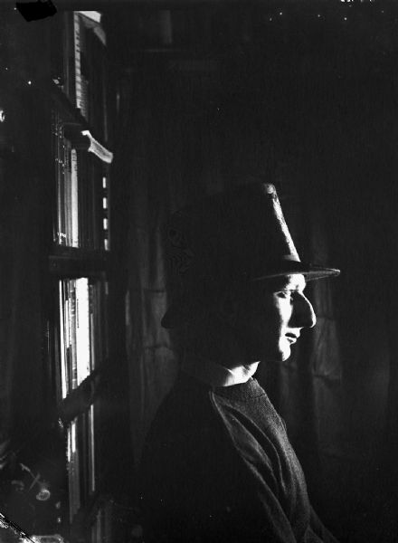 A man wearing a hat sits in profile in a darkened room with a bookshelf behind his back.