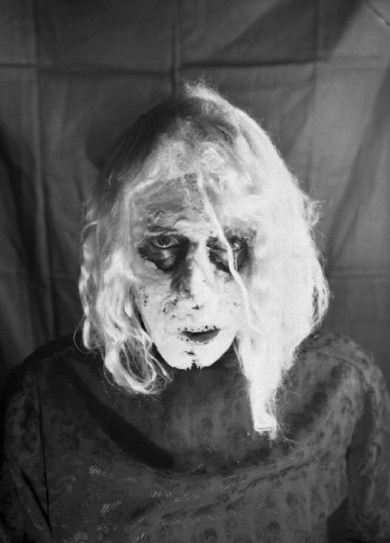 Robert Bloch in front of a drop cloth, wearing a thick layer of white monster makeup and a scary wig over a dark shirt.