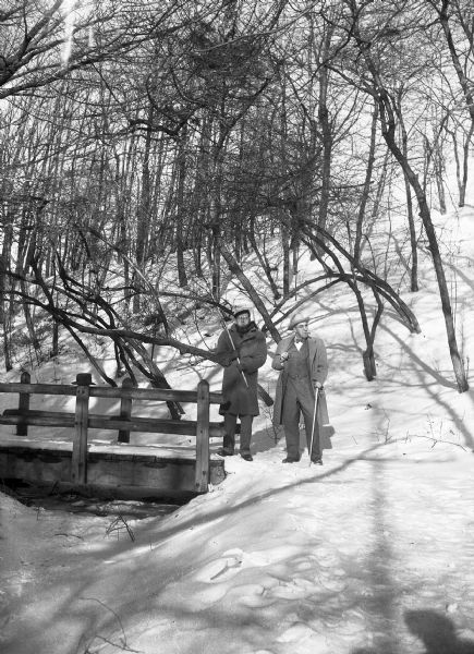 Harold Gauer and Robert Bloch stand near a wooden bridge, holding canes, in a wooded area blanketed in snow. There is a hat on top of one of the posts on the bridge.