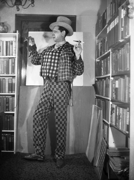 Robert Bloch, standing next to shelves of books, in front of a door covered with cardboard. He is wearing the gaudy costume of the King Geek for a photo shoot for a humor magazine.