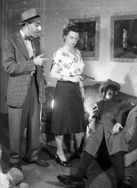 Harold Gauer, wearing makeup, a false nose and smoking a cigar, stands next to Alice Bedard, holding a revolver. They are standing over the body of Robert Bloch sitting on a couch, who is playing dead and wearing a hat, wig, and trench coat.