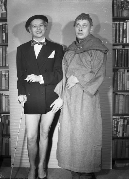 Robert Vail, dressed up as a Monk, subtly caresses the thigh of Angie Vail. Vais is smoking and wearing only a tuxedo jacket with a bow tie, hat, and shoes, and leaning on a cane.