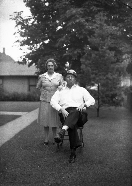 A man smoking a cigar and wearing a helmet sits on a chair on a front lawn with a woman, presumably his wife, standing behind him.