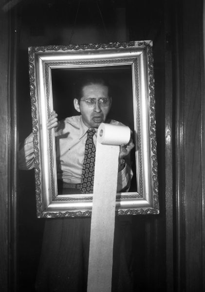 Robert Bloch standing in a doorway peers through an ornate picture frame holding a roll of toilet paper in front of his open mouth.