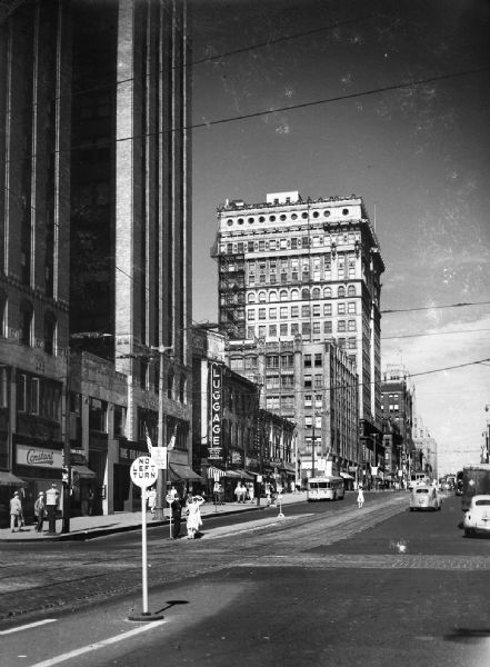 Looking east on Wisconsin Avenue at Water Steet with cars and pedestrians. Streetcars are running up and down the middle of the wide street. Skyscrapers and storefronts line the left side. The Wells Building is at the center of the photo.