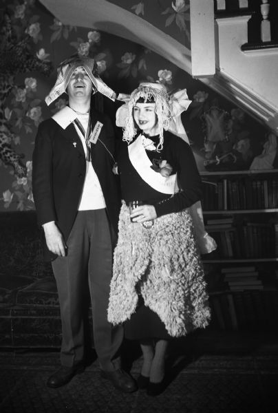 Max and Ruth Distenfeld stand under a stairway at a costume party. Max is wearing a hat made of newspaper and a badge that reads committee, while Ruth is wearing a bridal hat, a sash with obscured text, and what is likely a bath mat as a skirt.