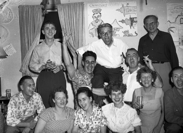 Large group of Harold Gauer's friends. In the front row from left to right are Bob Vail, Marion Bloch, Mary Jo Vonier, Angie Vail, an unidentified woman, and Robert Bloch. In the back row stands Sprague Vonier with his arm raised, helping to hold up a man on his shoulder. The other people are unidentified. On the wall behind them is a caricature of "Chinatown."
