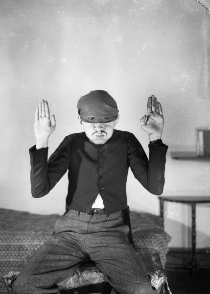 Robert Bloch sits on the edge of a bed with his hands up as in mock surrender wearing a hat pulled down over his eyes, but the hat has holes cut out of it so that his eyes are visible and he can see. Bloch is puffing out his cheeks and has a false moustache drawn on his upper lip.
