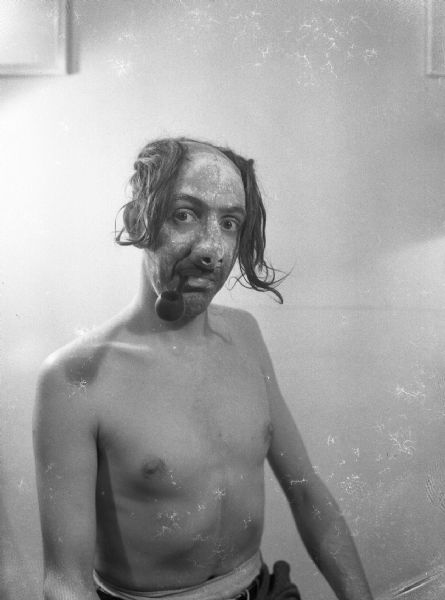 A shirtless Harold Gauer wearing elaborate facial make-up, a wig, and a prosthetic pig-like nose, and smoking a pipe.