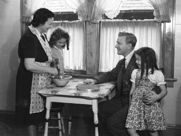 Milwaukee mayoral candidate (and future mayor) Carl Zeidler in election photograph sitting at kitchen table with a woman and two young children. There is a cake on the table and the woman is mixing ingredients in a bowl.