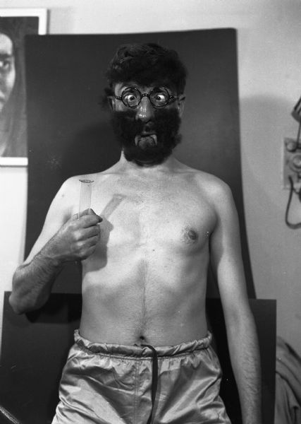 A shirtless Harold Gauer is wearing fake fangs, a beard, eyeglasses, a wig, and is holding a scientific test-tube.