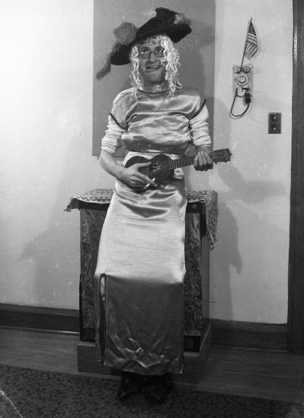 Robert Bloch wearing a dress, a blonde wig, eyeglasses and a feathered hat, strums a ukulele while holding a cigarette and sticking his tongue out. On the wall a small American flag is propped on the apartment intercom.