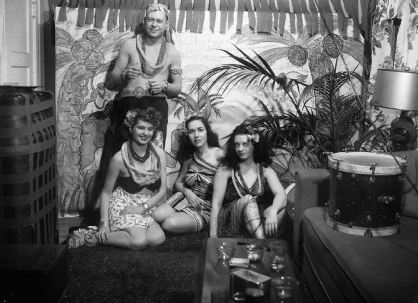 Robert and Angie Vail, Mary Jo Vonier, and Alice Bedard pose in front of a tropical backdrop wearing leis. A bass drum sits on a couch next to them.