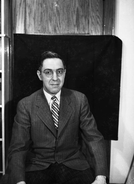A self-portrait of Harold Gauer wearing a jacket and tie, in front of a black backdrop.