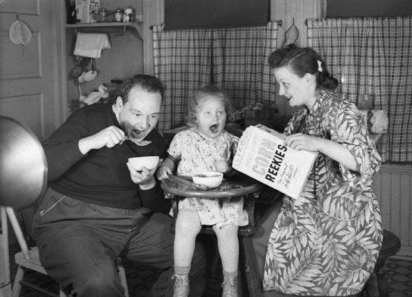An unidentified man, child, and woman sit around a kitchen table eating "Corn Reekies."